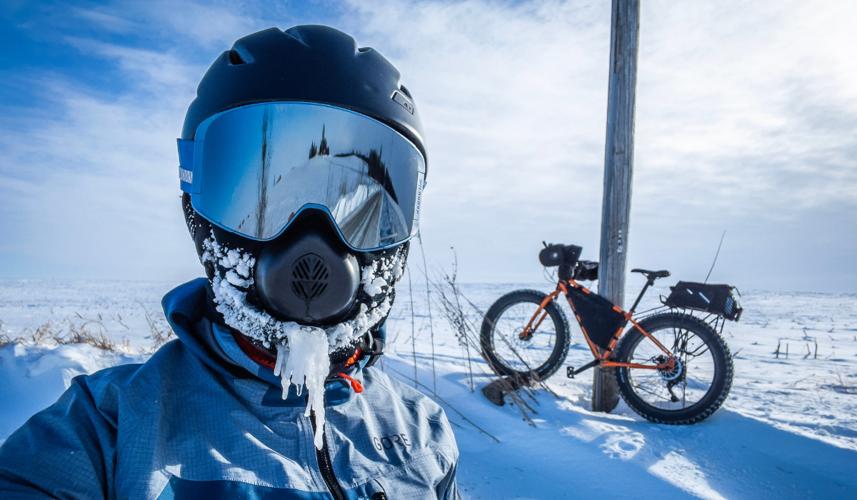 Person dressed in winter clothes and a bike helmet with a fat bike behind them in the snowy landscape